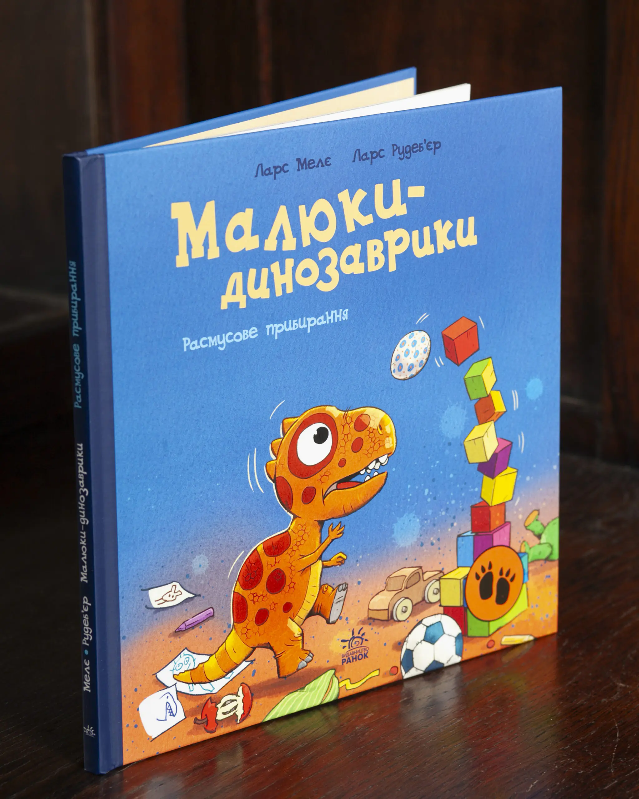 Малюки-динозаврики. Расмусове прибирання — передня обкладинка і корінець Малюки-динозаврики. Расмусове прибирання — книга Ларса Мелє на дерев'яному столі