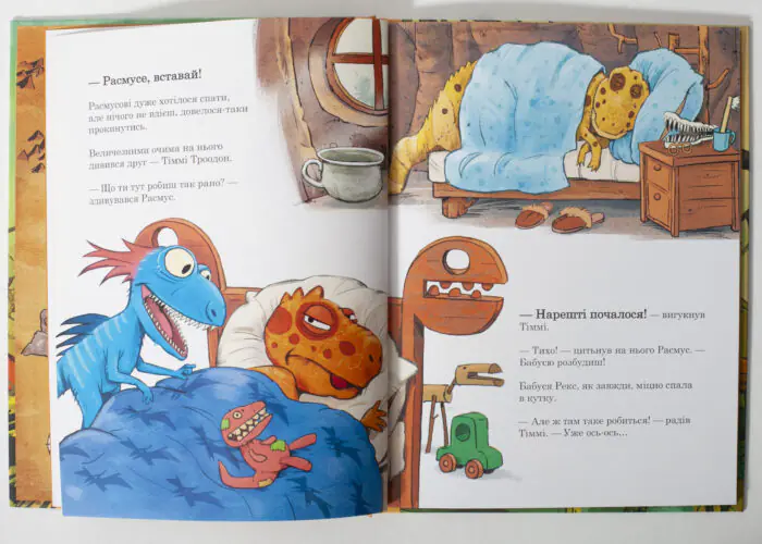 Two-page illustration: dinosaurs waking up in a cozy bedroom; a blue dinosaur sits up under a blue blanket while an orange spotted dinosaur sleeps in bed, with a dresser, mug, and glasses nearby.
